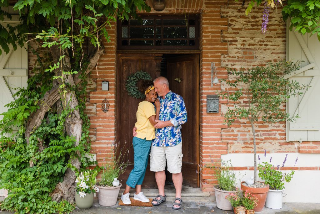 Image of a middle-aged couple happily embracing each other. They are standing in front of a brick building with a brown wooden double door surrounded by greenery