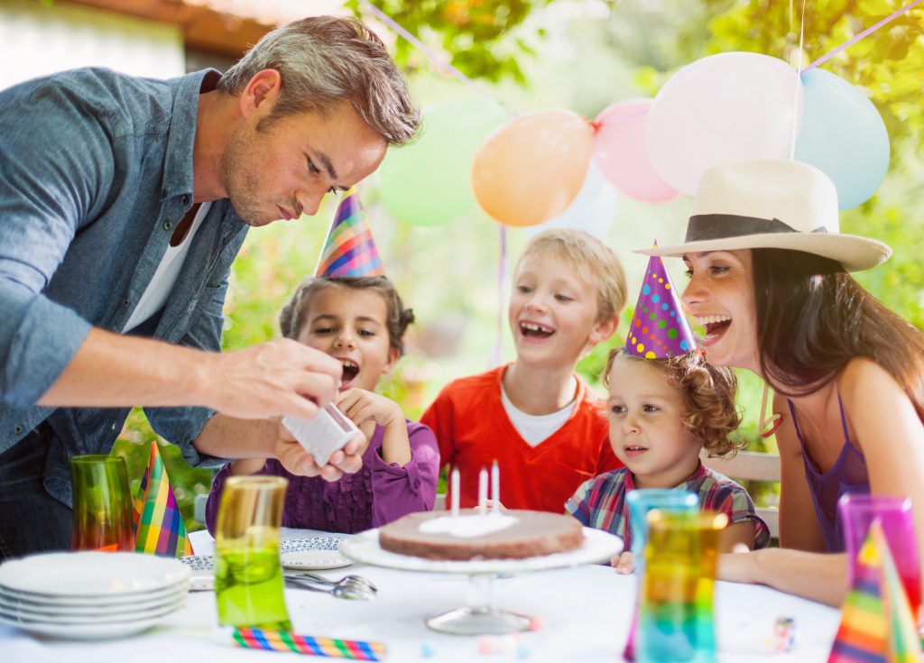 A family of 5 is colorfully celebrating a birthday outside their house with dessert, balloons, and party hats.