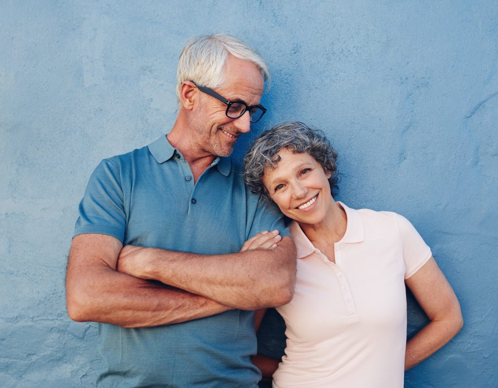 Image of a mature couple standing against a blue wall. The man is happily looking at the woman who is leaned up against him