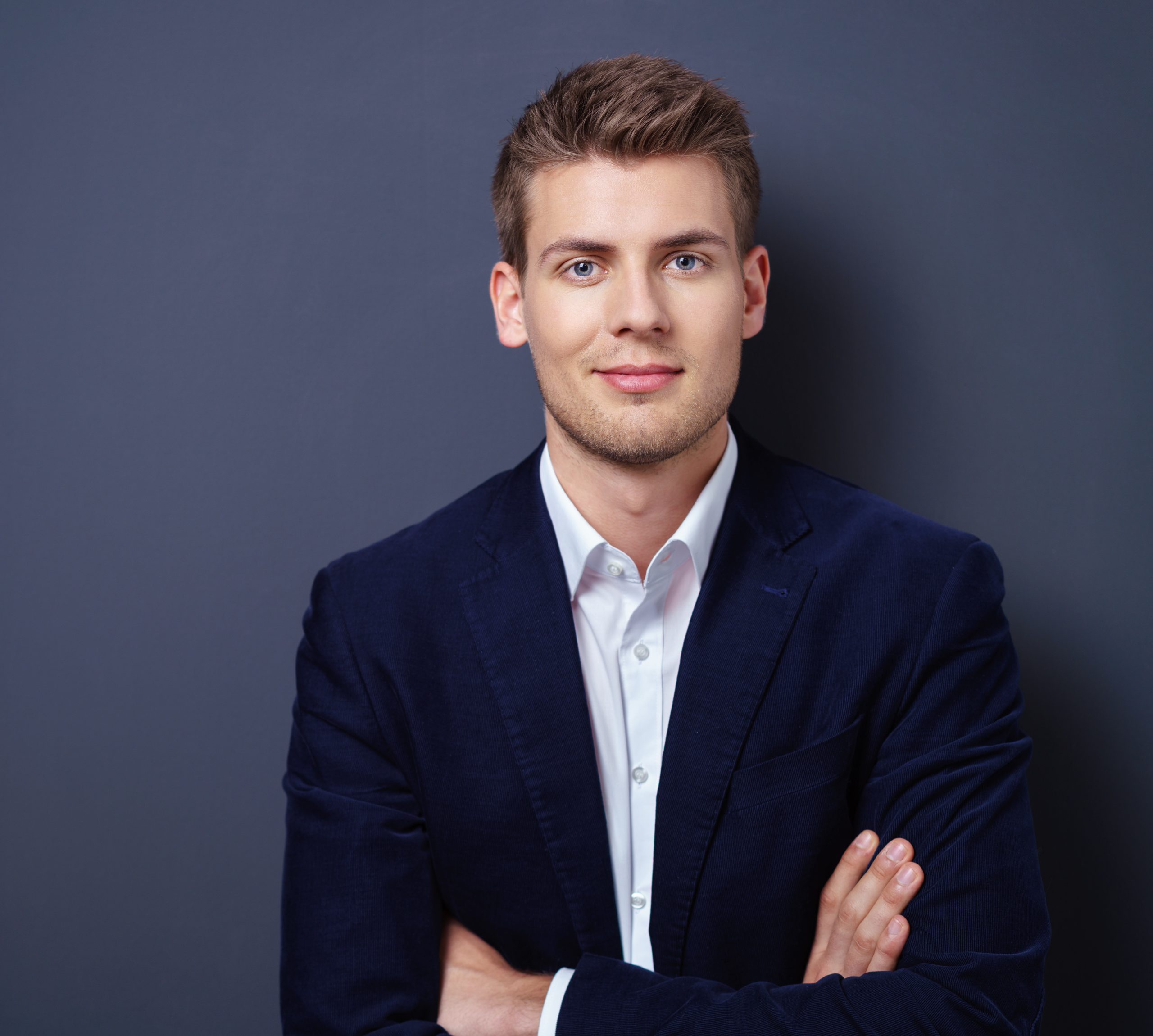 Young businessman in a dark blue coat standing with folded arms smiling at the camera in front of a dark background