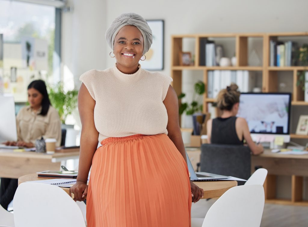 A smiling professional woman leans back against a table with paper and a computer on it. There are coworkers in the background working on Macs.