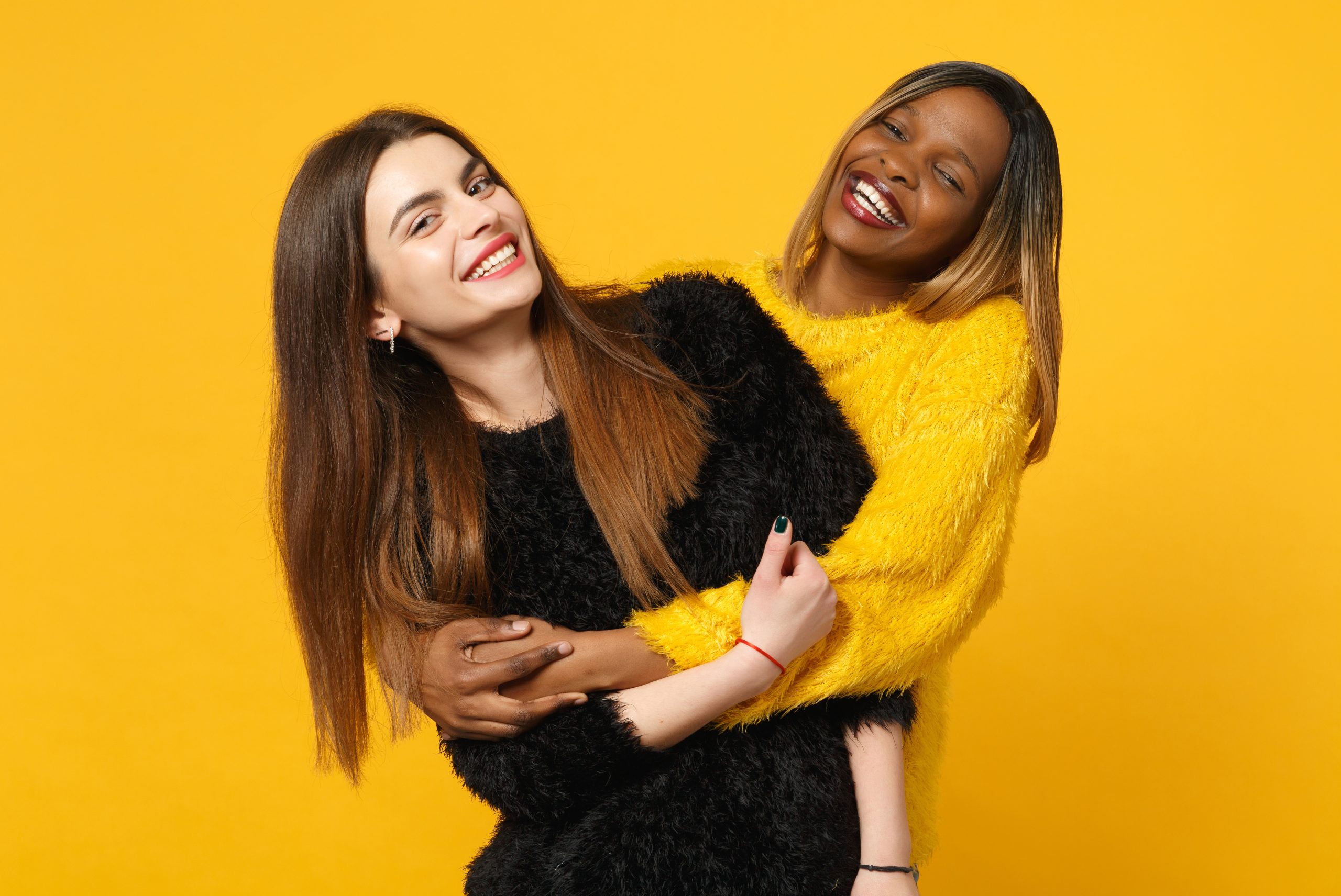 Two young women in black and yellow clothes hugging with a bright orange wall background