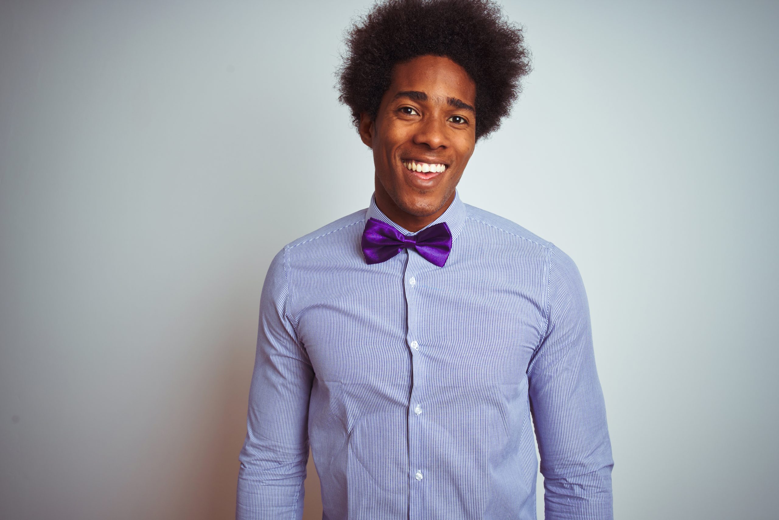 Young smiling man wearing a striped shirt and dark purple bowtie standing in front of a white wall