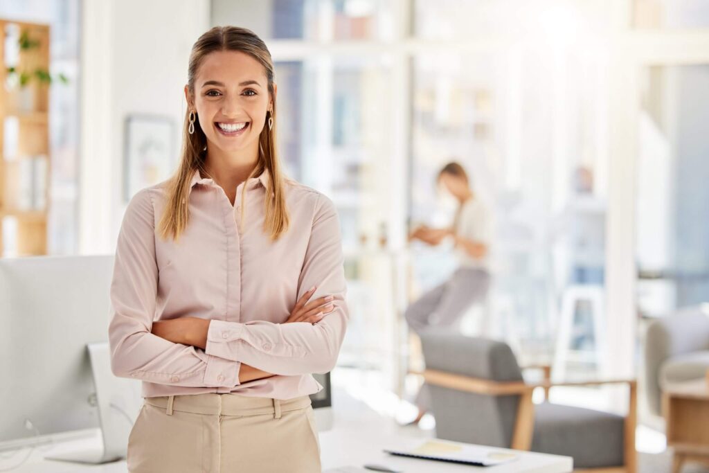 A professional looking woman is smiling with her arms crossed. The office and coworkers are blurred in the background.
