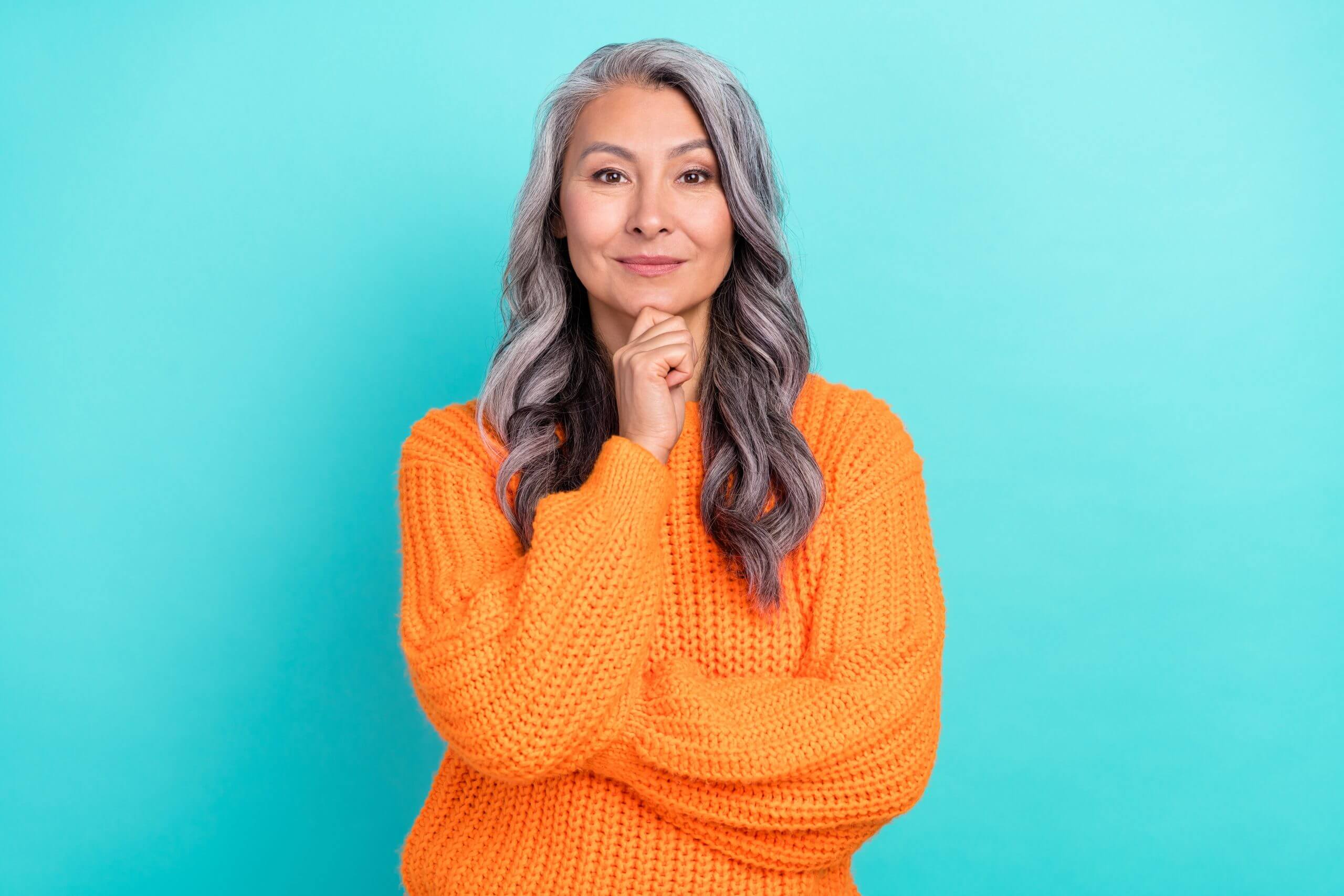 Portrait of a grey-haired woman with her hand on her chin standing in front of a teal turquoise background.